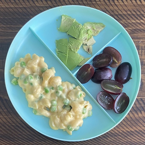 Plate showing toddler meal idea with mac & cheese with peas, avocado, and grapes.
